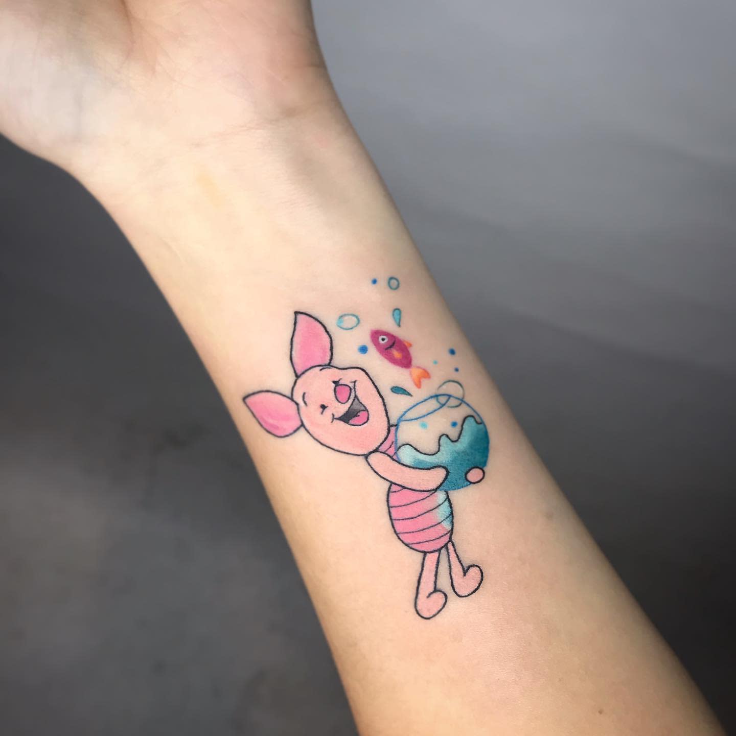 Tattoo of Piglet joyfully holding a fishbowl with a colorful fish on a person's forearm